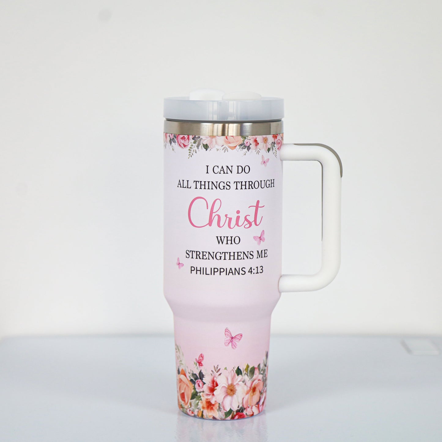 A pastel pink tumbler with a white handle and lid, featuring the Bible verse "I can do all things through Christ who strengthens me – Philippians 4:13," surrounded by floral and butterfly illustrations at the top and bottom.