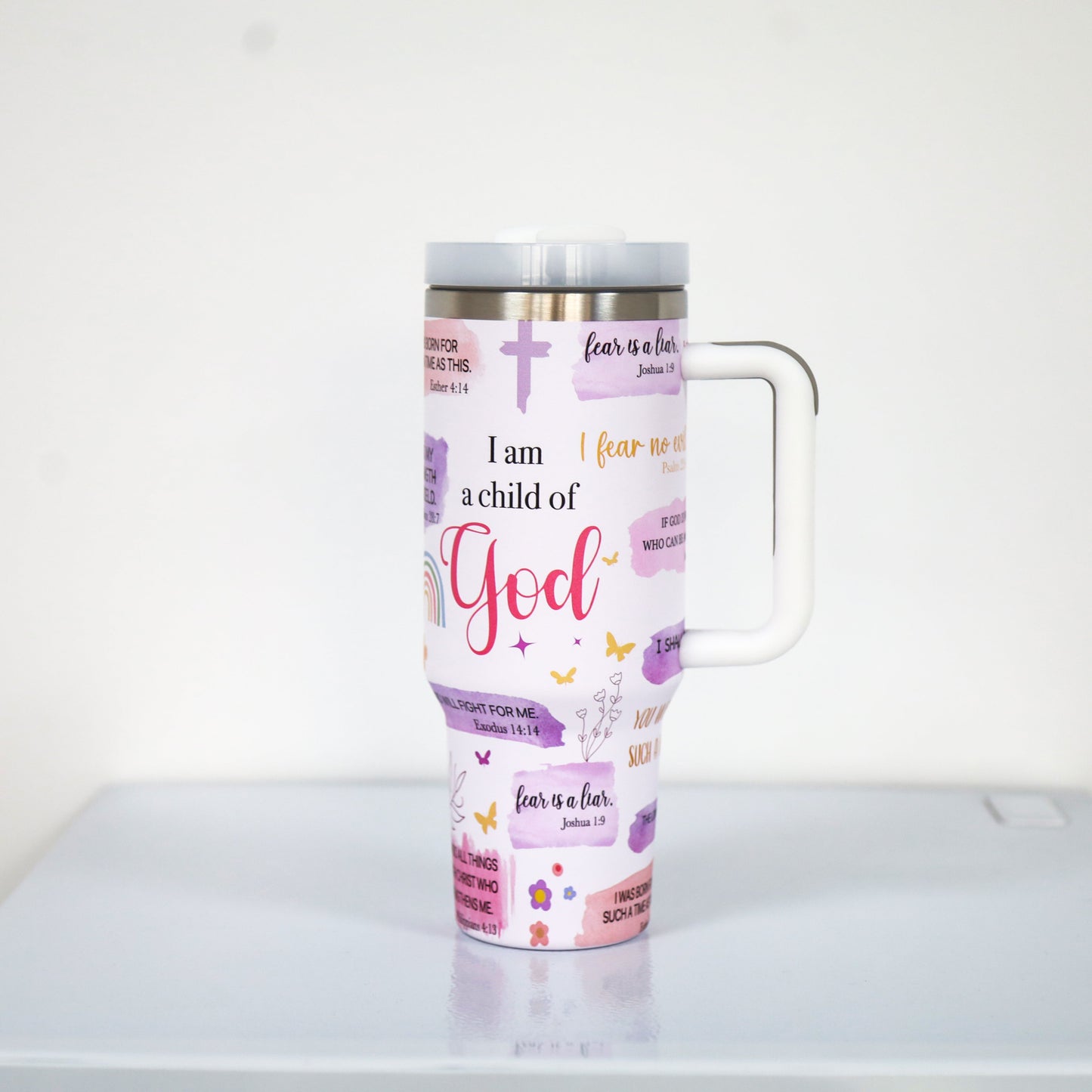 A pastel purple tumbler with a white handle and lid, featuring Christian faith-based quotes such as "I am a child of God" and "fear is a liar," along with crosses, rainbows, butterflies, and floral elements.