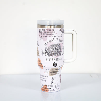 A pastel pink tumbler with a white handle and lid, decorated with Bible-themed affirmations and scriptures such as "My Daily Bible Affirmations," "All things are possible (Mark 9:23)," and floral illustrations surrounding a sketch of an open Bible.