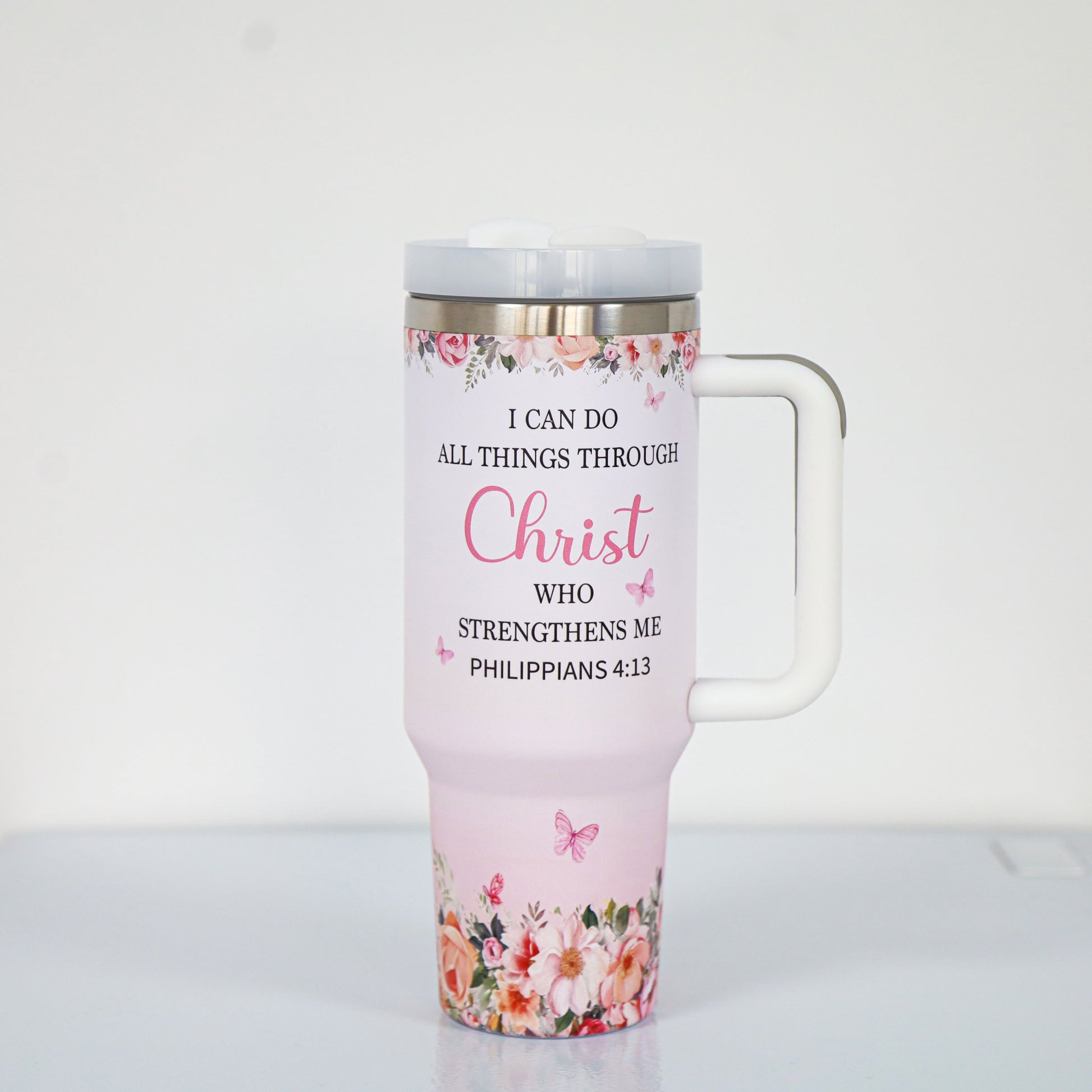 A pastel pink tumbler with a white handle and lid, featuring the Bible verse "I can do all things through Christ who strengthens me – Philippians 4:13," surrounded by floral and butterfly illustrations at the top and bottom.