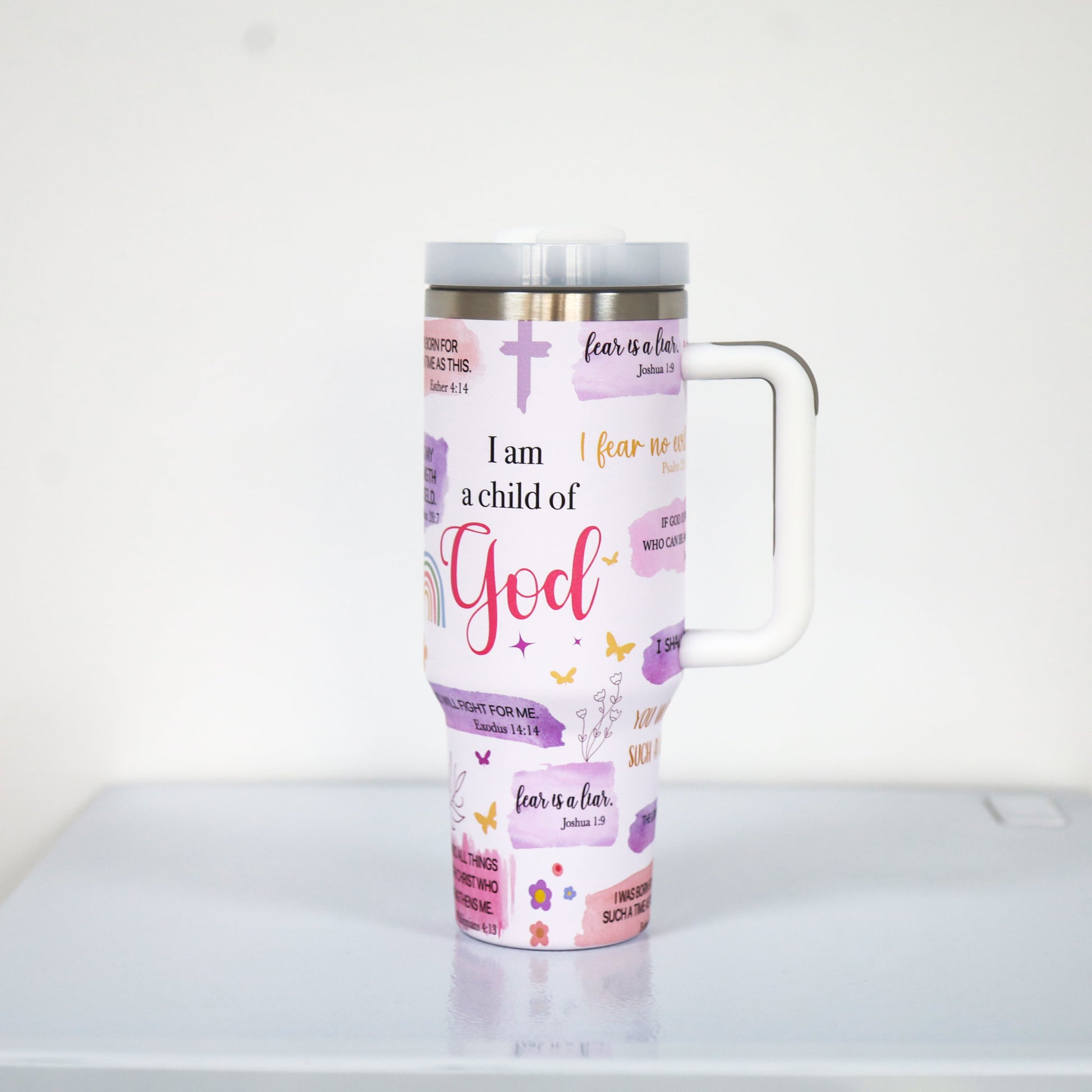 A pastel purple tumbler with a white handle and lid, featuring Christian faith-based quotes such as "I am a child of God" and "fear is a liar," along with crosses, rainbows, butterflies, and floral elements.